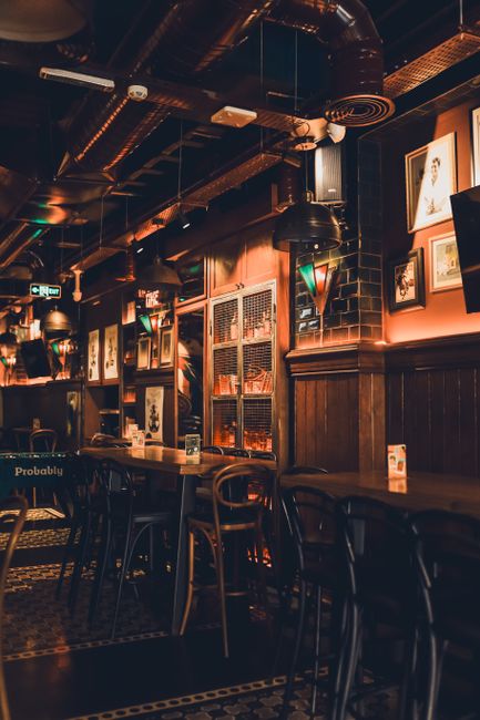 Interior view of a dimly lit pub with wooden paneling on the walls and a patterned tiled floor. The ceiling features exposed black ducts and industrial-style pendant lights casting warm light. Framed 