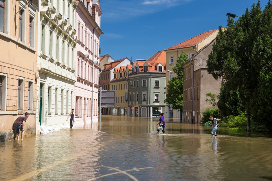 A wide view of a river in Brompton SW3 showing a partially submerged streetlight post in the foreground with water reaching up to its base. Across the river, a cityscape features a historic church wit